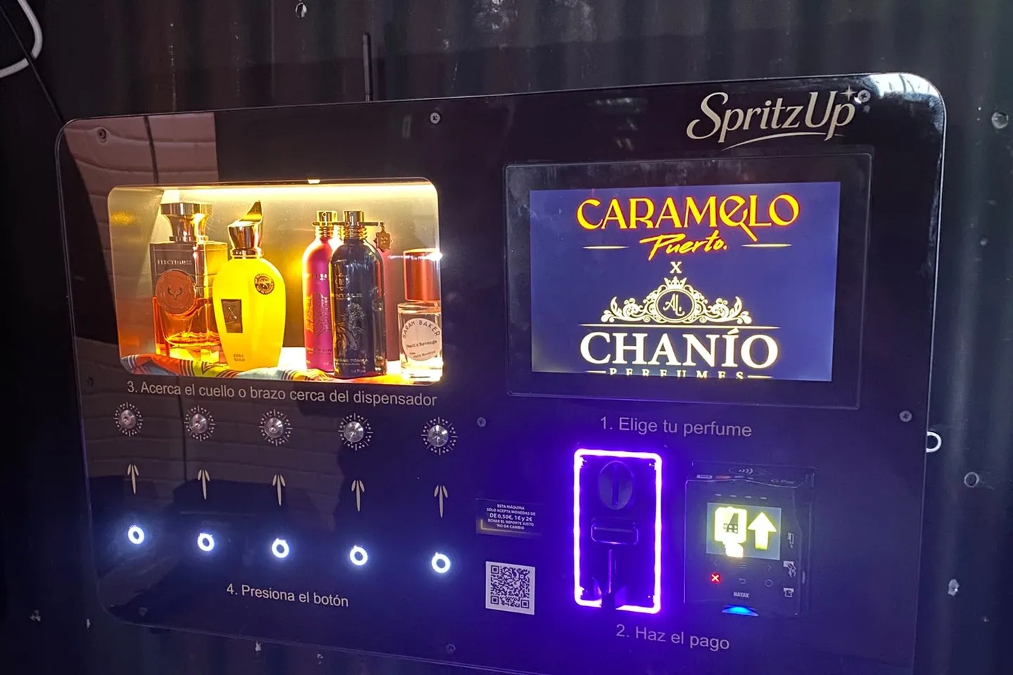 A SpritzUp perfume vending machine mounted on a corrugated wall at the venue at night: the perfume shelf is lit gold, the touchscreen is running the venue’s advert, and the coin slot glows violet.